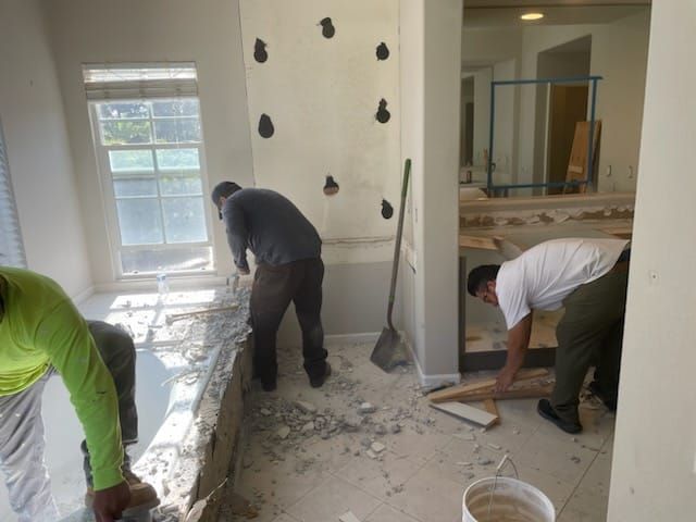Three workers demolish a room. One uses a tool on a window sill, another on the wall, and another on the floor.