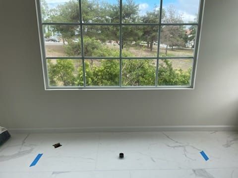 Room with large window overlooking trees, white tile floor, blue tape, and black object.