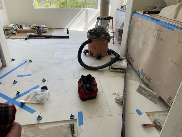 Floor tiling in progress; white tiles, blue tape, vacuum, and tools are visible in a room.