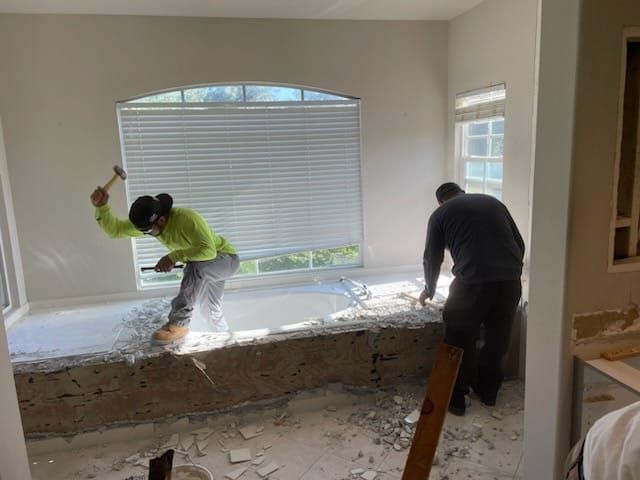 Two workers removing a bathtub surround. One swings a hammer, the other uses a tool. The bathroom has a window.