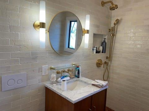 Bathroom with round mirror, gold fixtures, marble countertop, and off-white brick-like tile.