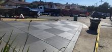 Concrete checkered sidewalk next to a curved curb. Trucks and buildings in the background on a sunny day.