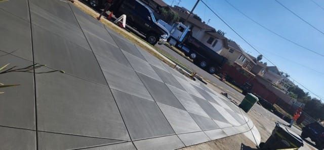 View of a driveway with checkerboard-patterned concrete. A truck and buildings are visible in the background on a sunny day.
