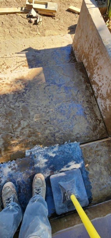 Person cleaning a concrete surface with a large blue sponge attached to a yellow pole.