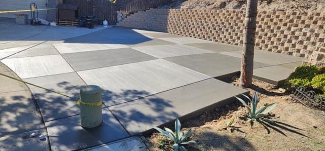 A newly paved concrete surface with a checkered pattern, next to a retaining wall and vegetation.