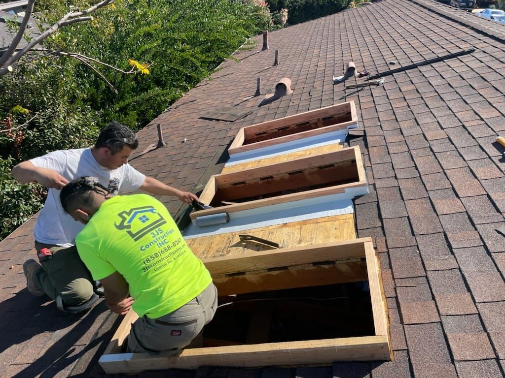 Two workers on a rooftop, installing skylights. Brown shingles, open wooden frames, sunny outdoor setting.