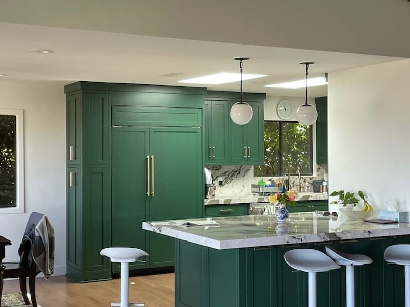 Green kitchen with island, cabinets, and built-in refrigerator. White countertops, pendant lights, and bar stools.