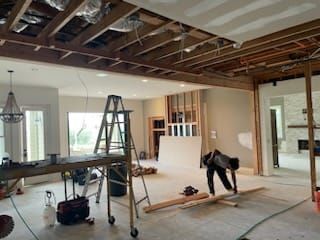 Interior renovation: room with exposed ceiling beams, drywall, ladder, tools, and a person working.