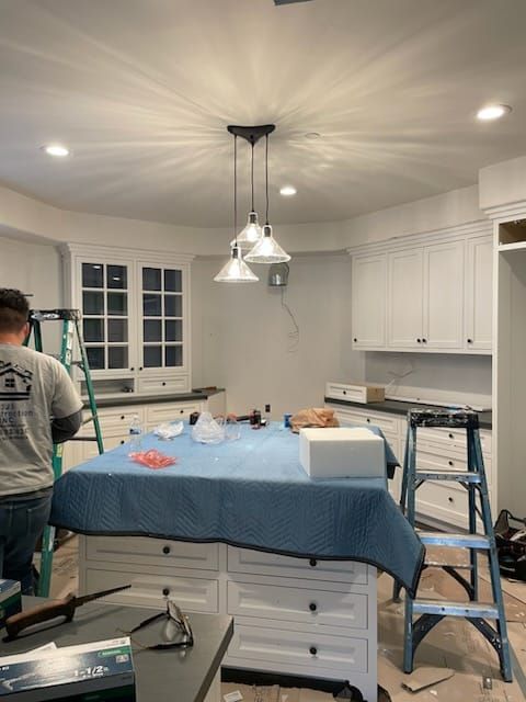 Kitchen under construction; cabinets, island, new pendant lights, person on ladder, tools on island.