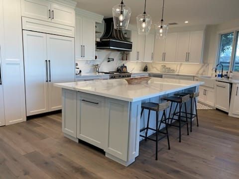 Modern white kitchen with island, marble countertops, pendant lights, and wooden floor.