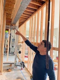 Man points to a beam in a framed wall under construction; interior setting with a worker on a lift.