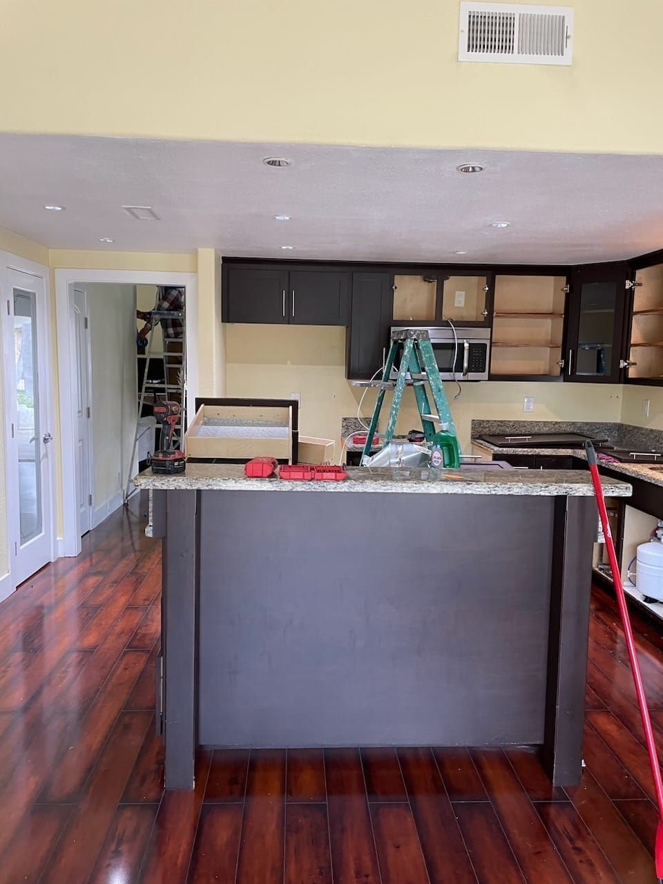 Kitchen undergoing renovation with dark cabinets, island, and ladder. Yellow walls and hardwood floors.