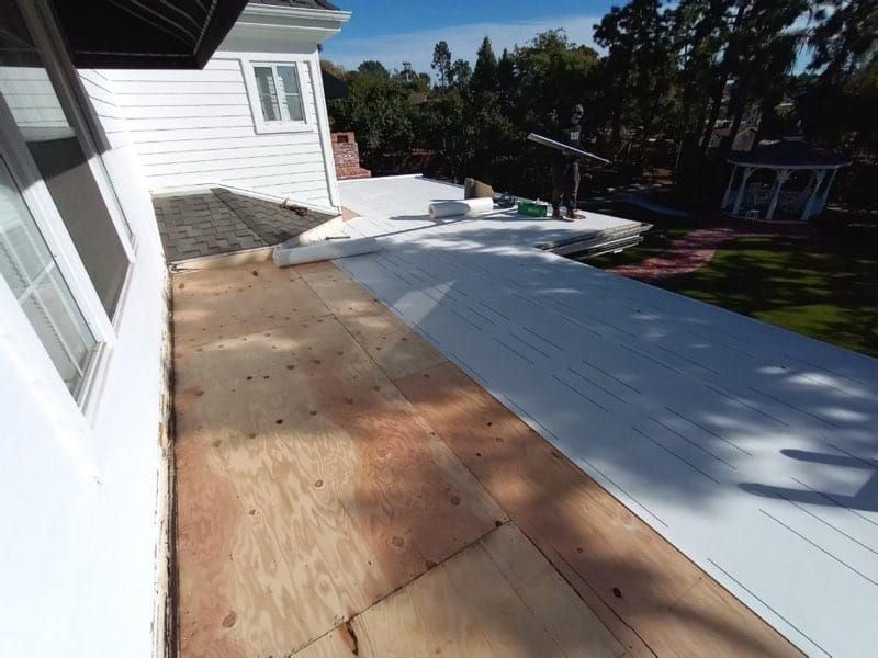 Exterior view of a house with a partially completed flat roof. White siding, wood deck, and green lawn.