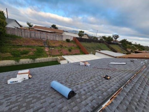 Rooftop with gray shingles; construction materials on the roof. Fenced hill and cloudy sky in background.
