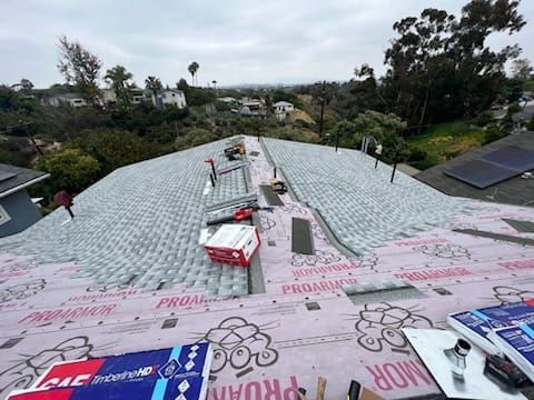 Roof being worked on, with materials and tools visible; houses and trees in the background under an overcast sky.