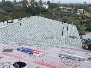 Roof with gray shingles, tools, and materials; houses and trees in background.