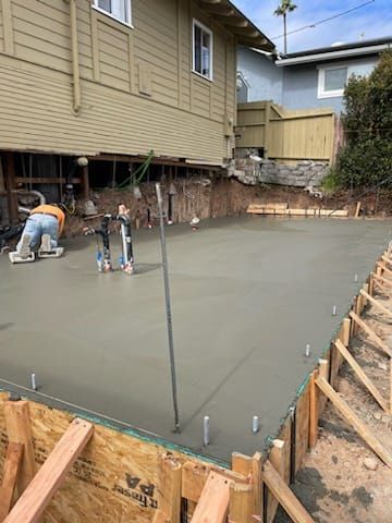 Construction workers pouring and smoothing concrete for a foundation, wooden forms in foreground, under a house.