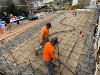 Workers pouring concrete on a grid of rebar. Two men in orange shirts use a hose. Other workers in background.