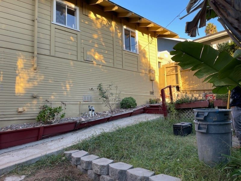 Side of a yellow house with a ramp and flower boxes; small yard with trash can and greenery.