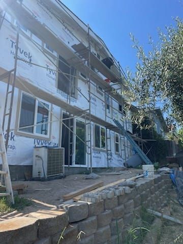 Two-story house under construction, covered in Tyvek wrap, with scaffolding. Retaining wall in the foreground.