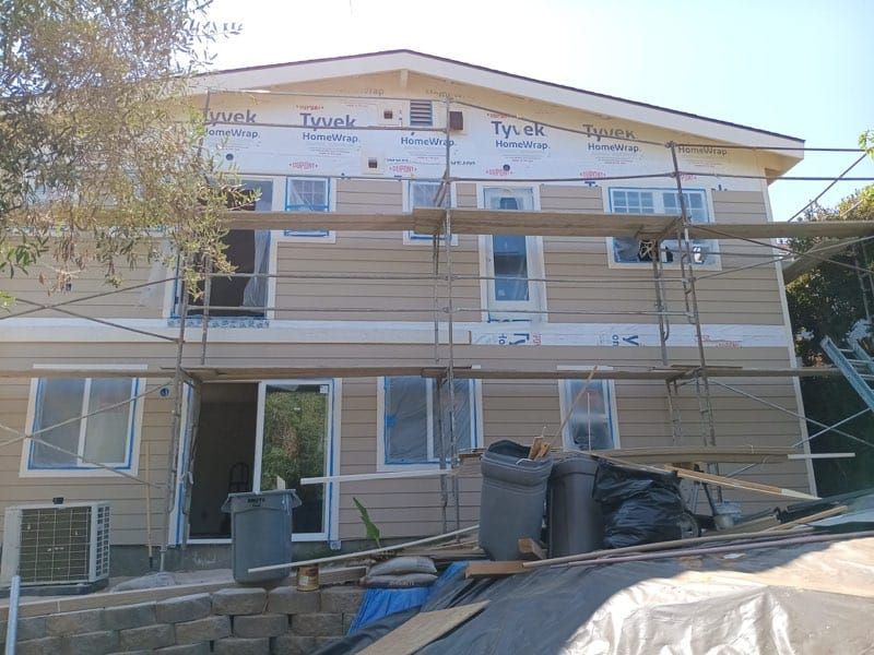Two-story house under construction, tan siding, scaffolding, white Tyvek wrap, windows, sliding glass door.