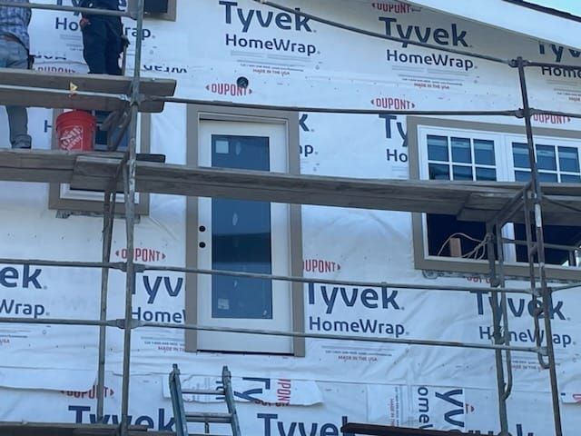 Exterior of a house under construction; Tyvek HomeWrap covering the walls; scaffolding surrounds a door and window.