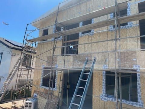 Building under construction; scaffolding surrounds a two-story structure with exposed wooden panels, blue sky overhead.