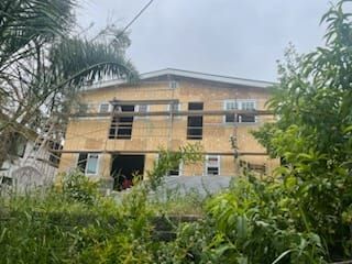 Multi-story house under construction, framed with exposed wood and scaffolding, surrounded by greenery. Overcast sky.
