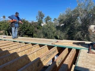 Construction workers installing roofing on a sunny day. OSB and rafters visible.