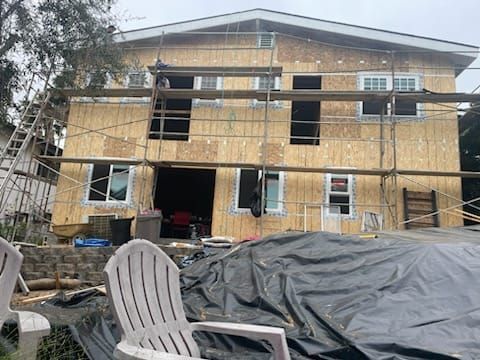 Two-story house under construction, sheathed in plywood. Scaffolding, windows, and chairs are visible.