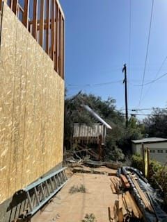 Construction site with wooden framing, plywood siding, and debris, with a tree in the background.