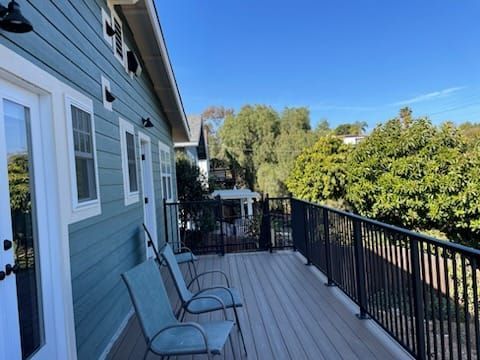 Teal house with a deck. Two chairs sit on the deck, which overlooks a yard with trees under a blue sky.