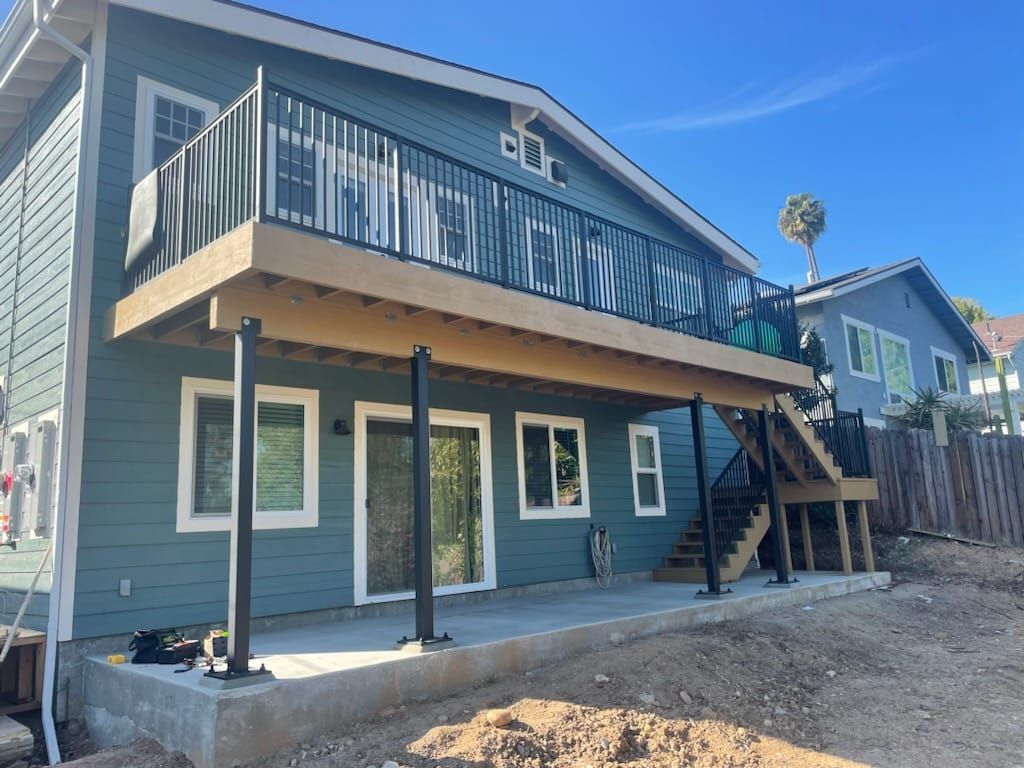 A two-story blue house with a raised wooden deck supported by black poles. Concrete patio below.