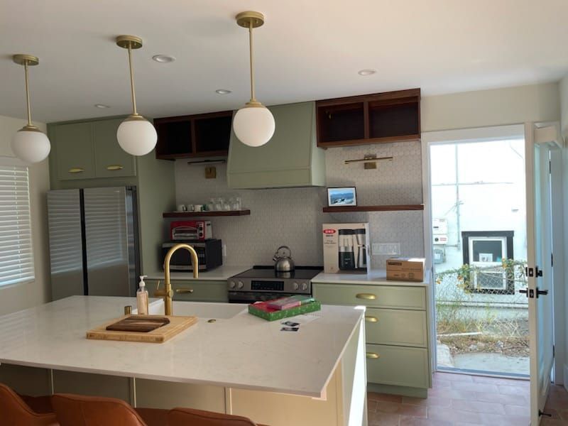 A light green kitchen with white countertops and gold fixtures. Includes an island, open door, and globe pendant lights.