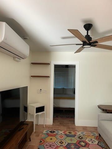 Living room with white walls, TV, AC unit, shelves, door to bedroom with bed, and ceiling fan.
