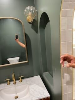 Green bathroom with marble vanity, gold fixtures, arched mirror, niche, and shell sconce.