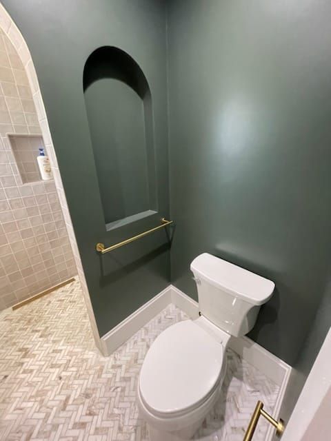 Small bathroom with a toilet and a recessed shelf. Dark green walls, gold fixtures, and patterned floor tile.