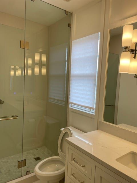 Bathroom with glass shower, toilet, white vanity, and window with shade.