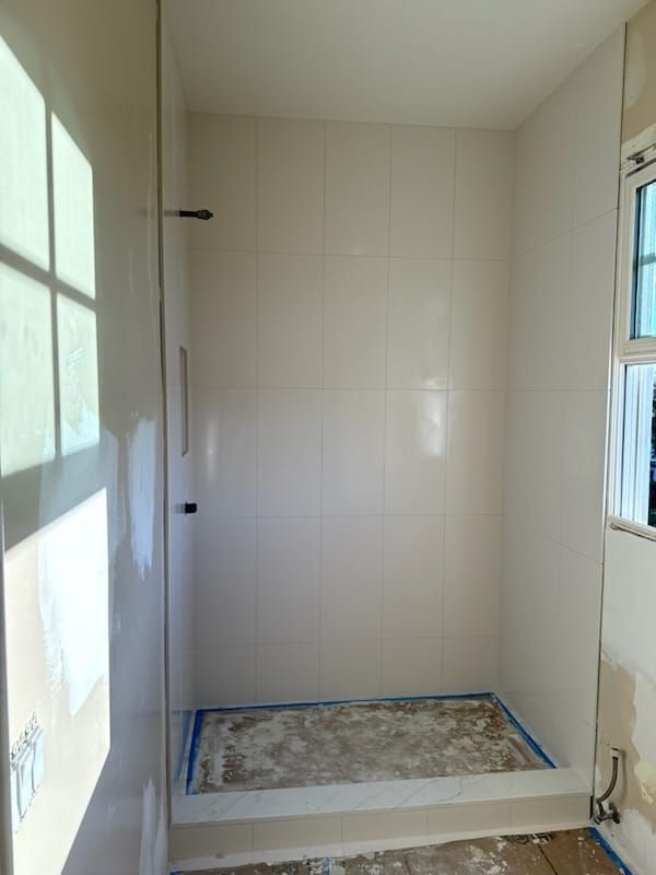 Empty shower stall with off-white tiles, shower head, and blue tape along the floor's edge.