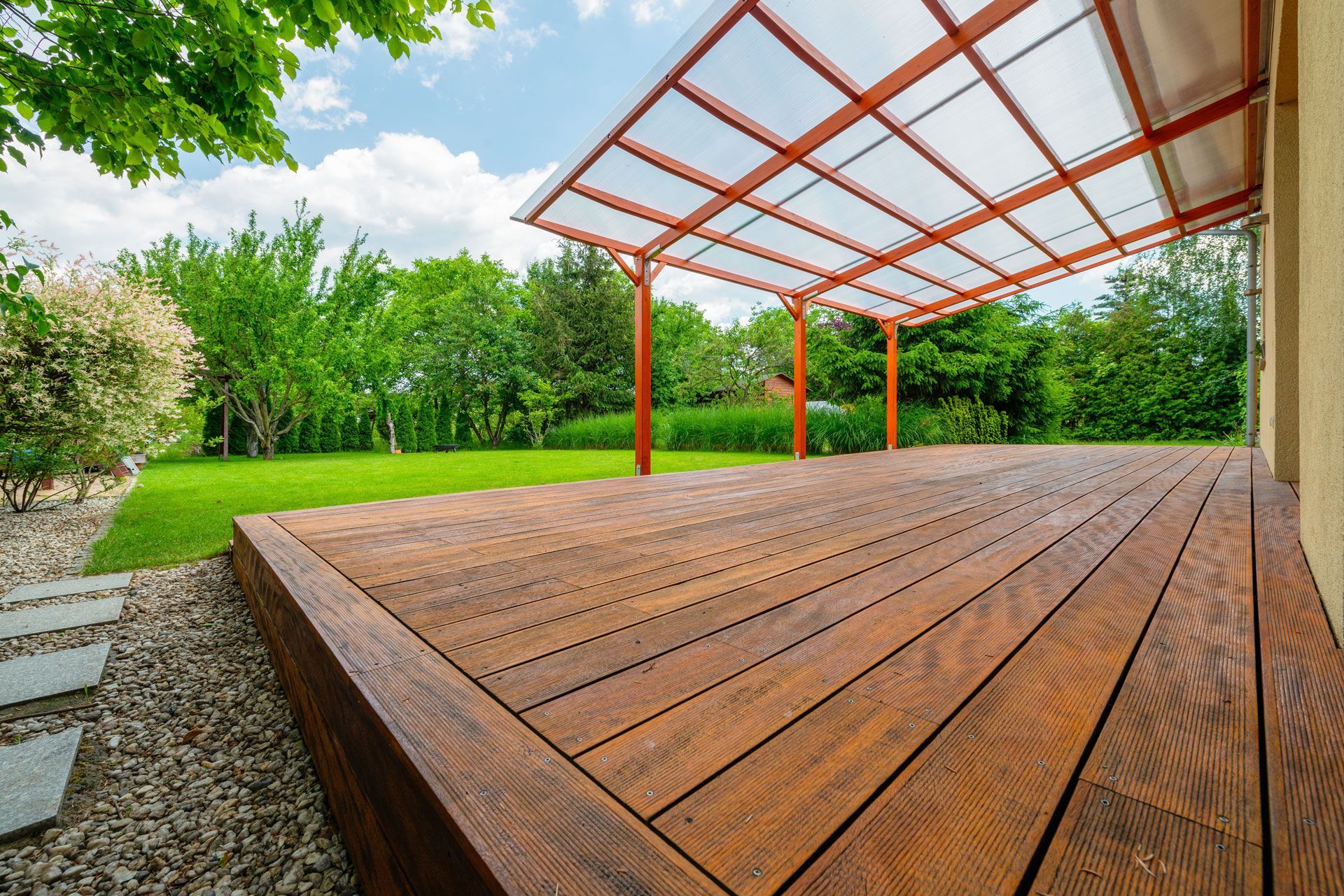 Wooden deck with brown pergola and clear roof, overlooking a green lawn.
