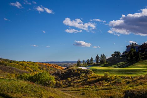 Golf course with houses on a hill against a blue sky. Green grass, trees, and buildings visible.