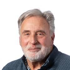 Persoon in gele coltrui, peinzend naar beneden kijkend, hand bij het gezicht, tegen een oranje achtergrond.