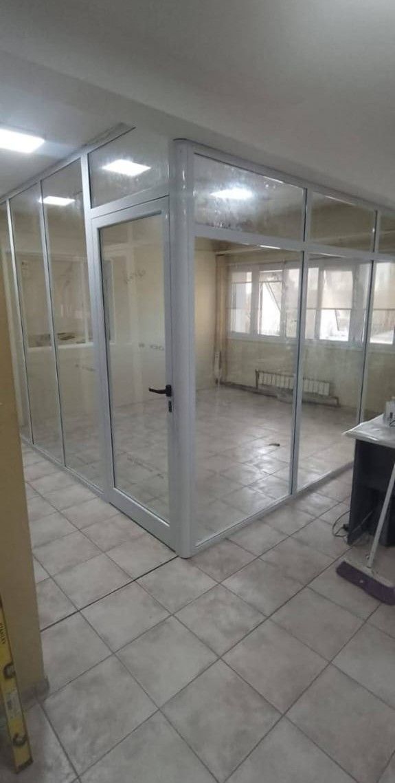 Cubículo de oficina con paredes de vidrio y puerta, ubicado dentro de una habitación más grande con piso de baldosas y ventanas.