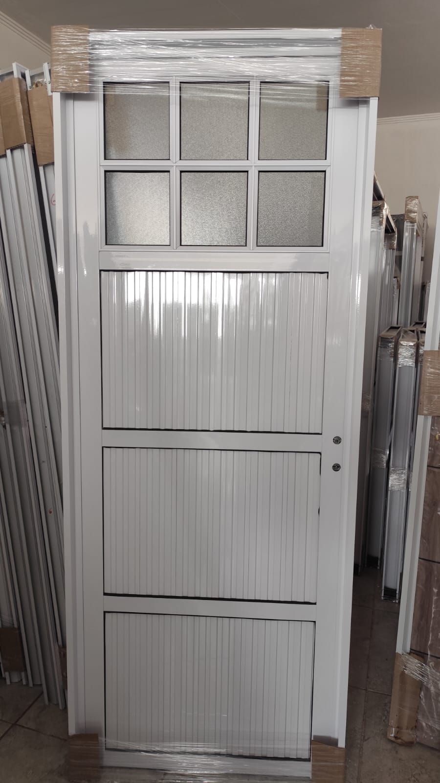 Puerta de metal blanca con panel de vidrio y líneas verticales, colocada en el interior de una puerta.