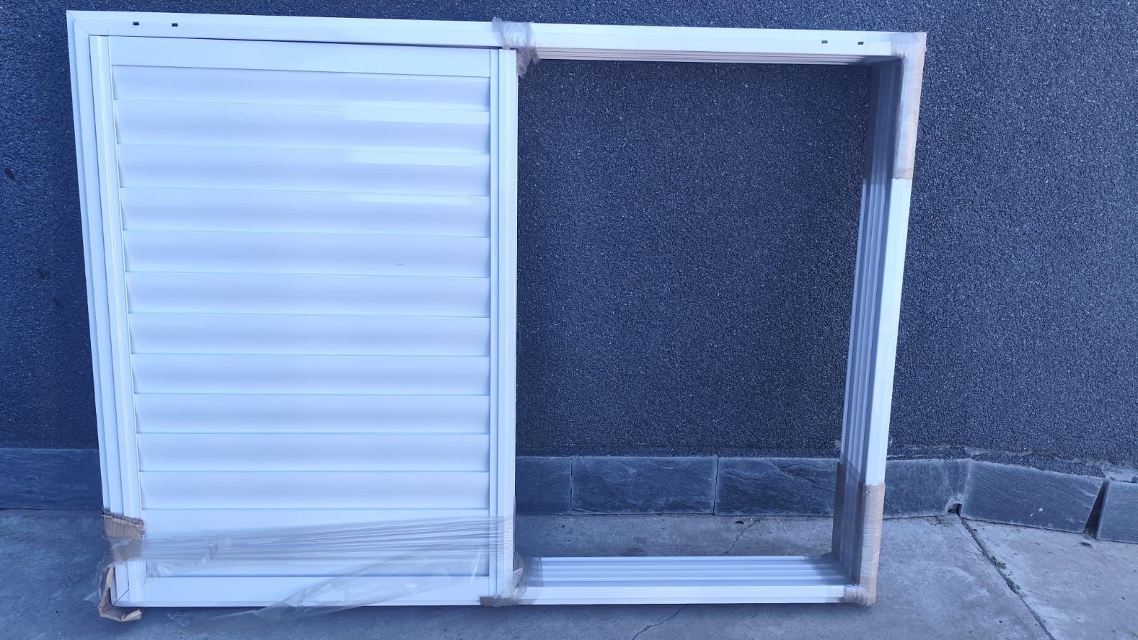 Panel de ventana con lamas horizontales de color blanco que cubre parcialmente un marco y descansa sobre una superficie gris texturizada.