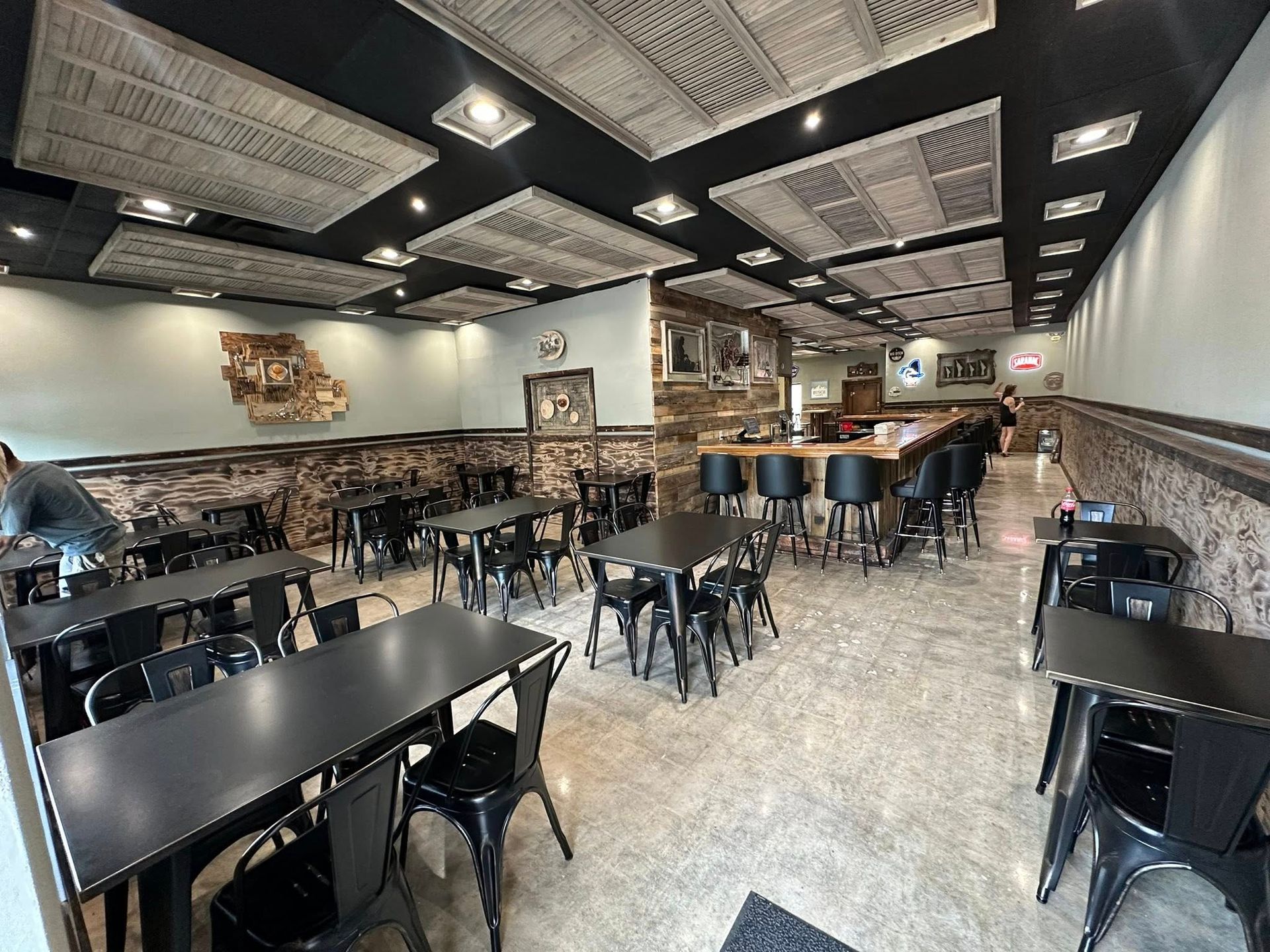 Interior view of a restaurant. Black tables and chairs are arranged in rows. A bar and decorative ceiling tiles are visible.