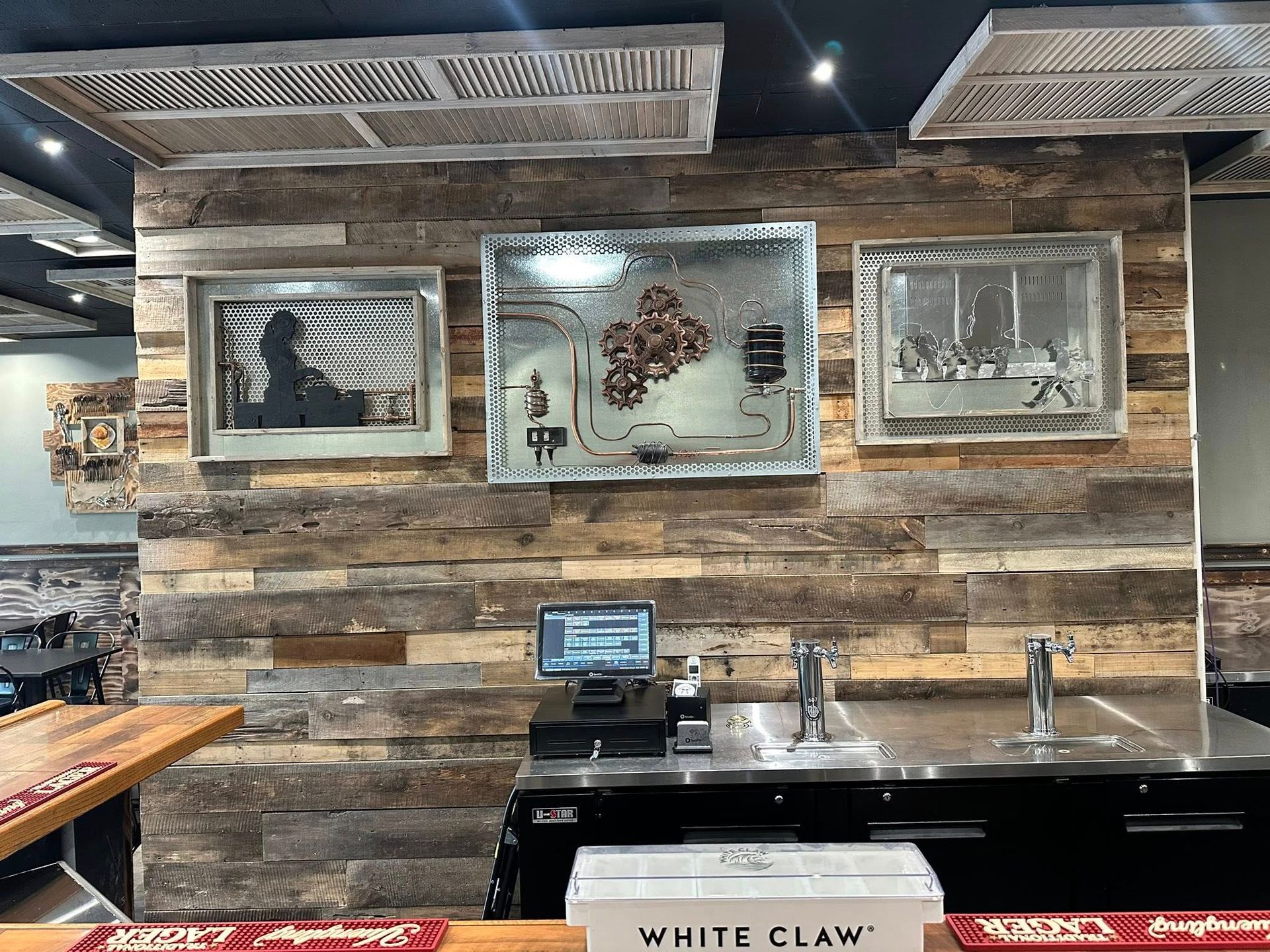 Rustic bar with wood paneling, artwork, and taps. A point-of-sale system sits on the stainless steel counter.