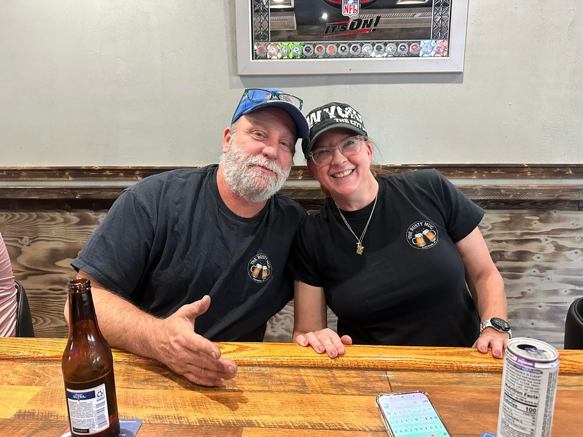 Two people smiling at a bar. Man with beard, cap. Woman with cap, watch. Beer bottle, can, wood bar.