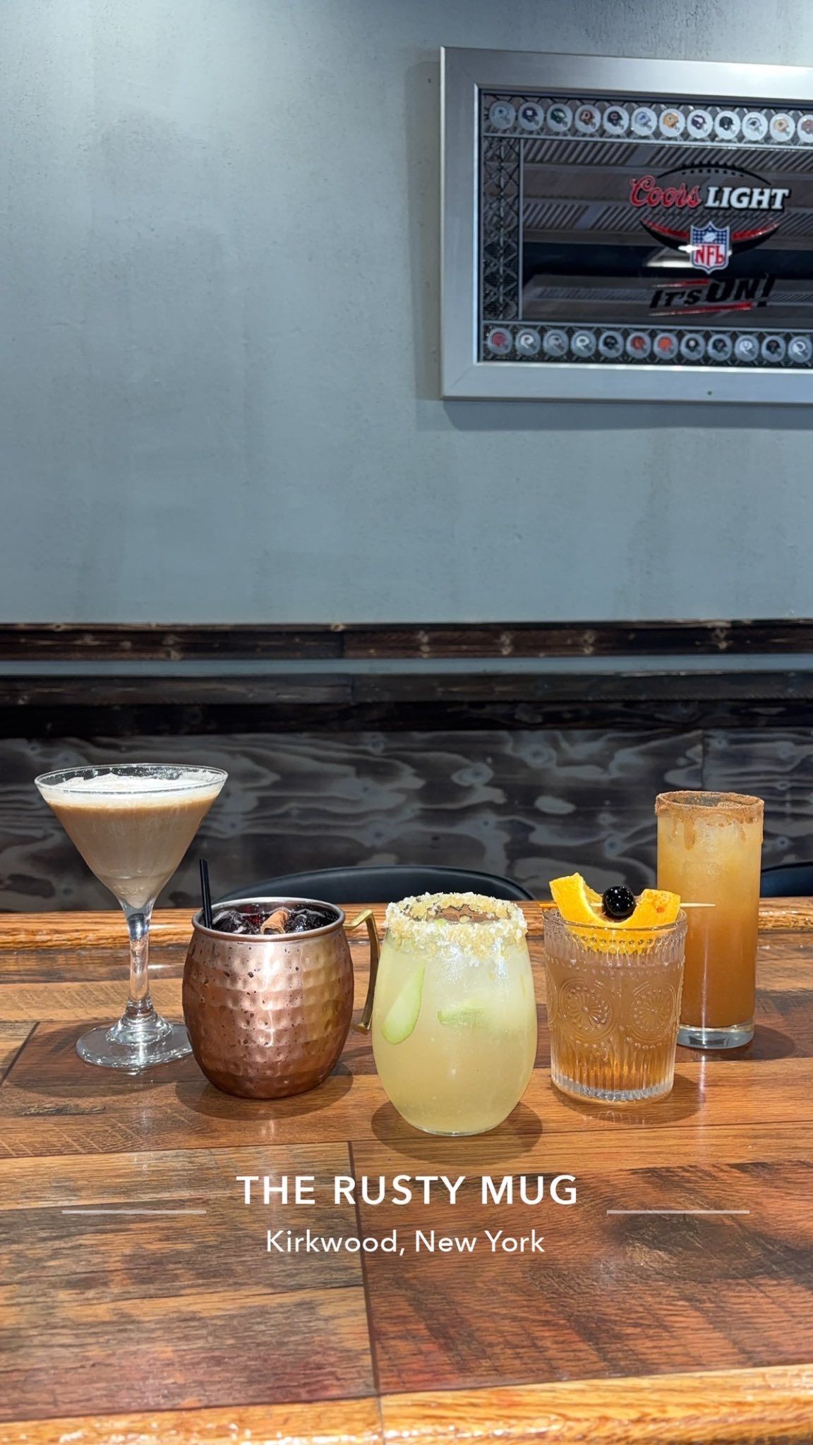 Cocktails on a wooden bar at The Rusty Mug, Katonah, New York. Drinks are in various glasses, with a metallic wall hanging.