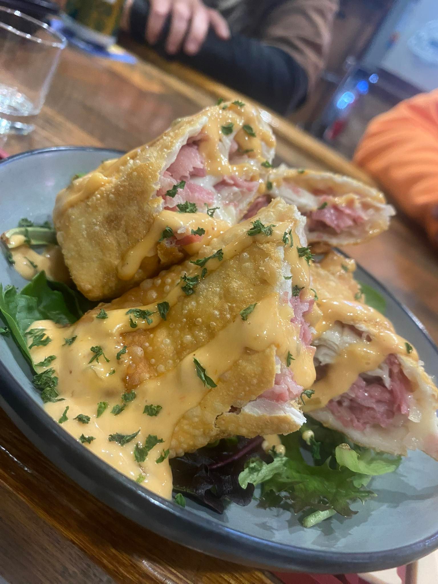 Fried food rolls, likely sandwiches, on a bed of greens, drizzled with sauce and parsley, in a blue bowl.